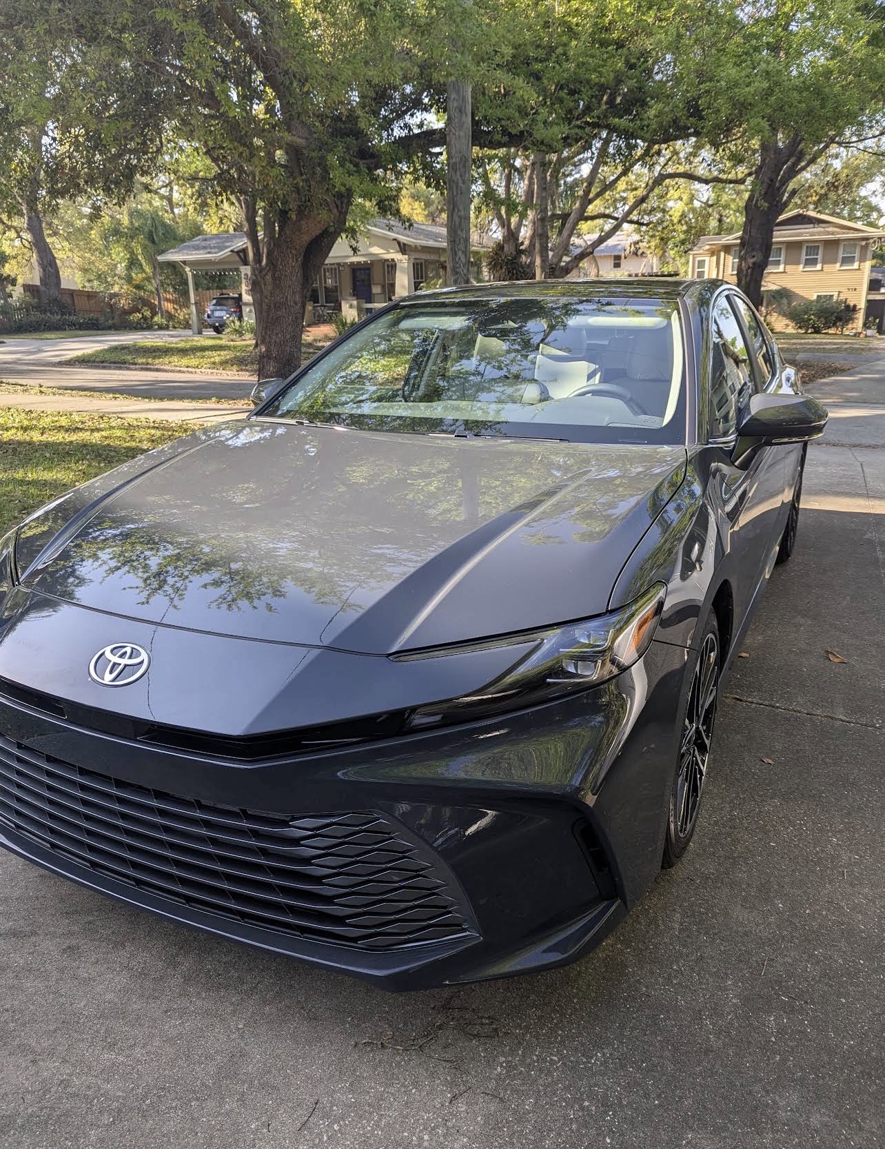 Freshly detailed black Toyota Camry with a deep, mirror-like gloss finish reflecting the trees overhead